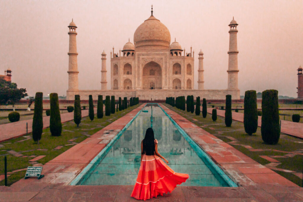 A landscape photo of myself standing by the ornate reflective pool, one of the best places to photograph the Taj Mahal from, at sunrise. The Taj Mahal has a golden cast due to the morning rays and there are perfectly manicured gardens all around the turquoise pool. 